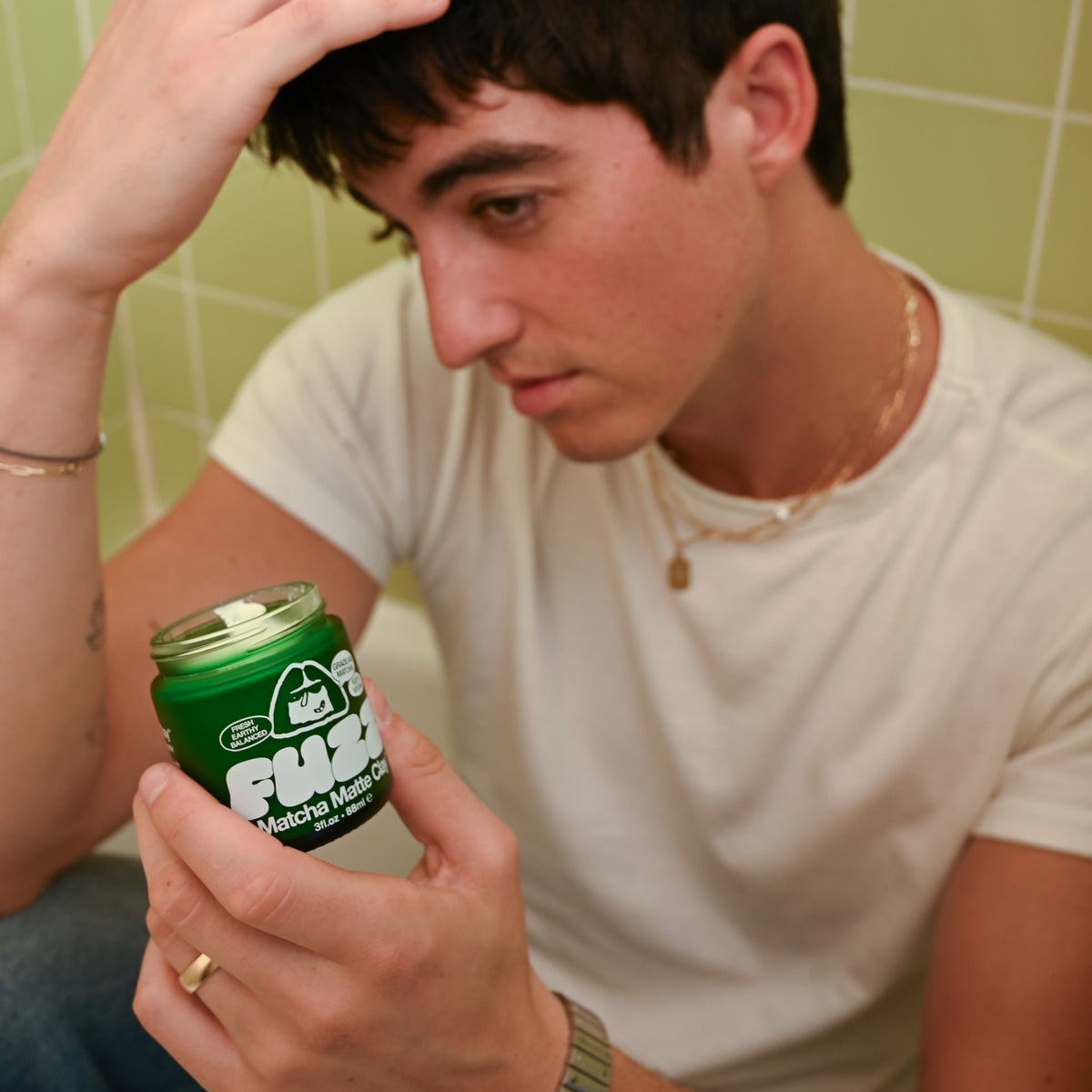 Person holding a green jar of hair product labeled FUZZ in a bathroom setting